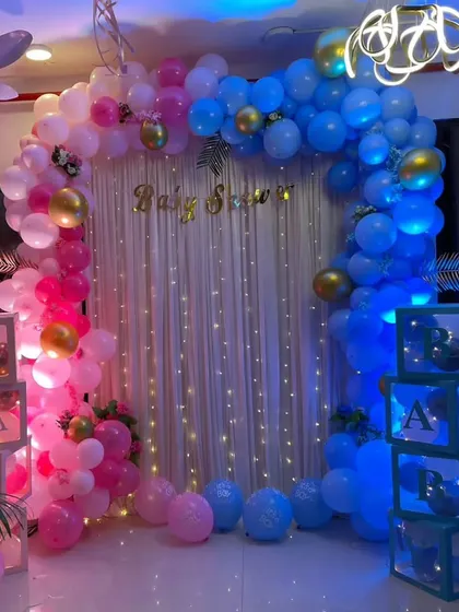 A gender reveal baby shower with a split pink and blue balloon arch under a fairy light curtain.