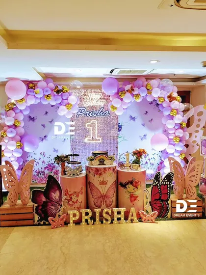 A stunning butterfly theme for Prisha's first birthday, featuring a sequin backdrop and a purple and gold balloon arch. The large butterfly props and 'BABY' blocks complete the glamorous look.