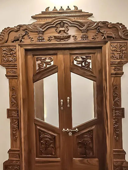A close-up of a beautifully carved pooja room door, showing the intricate details and the combination of solid wood and glass panels.