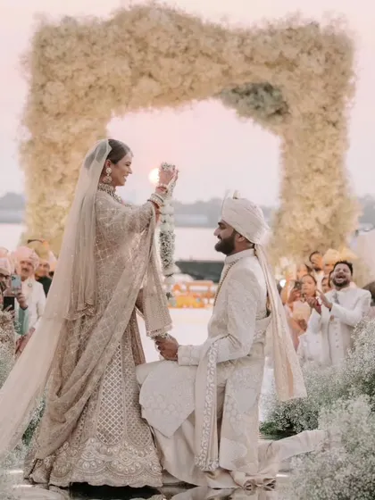 The bride places the varmala on her kneeling groom, a moment of playful tradition and deep affection. The sunset over the Kochi backwaters provides a stunning, natural backdrop to their all-white floral mandap.
