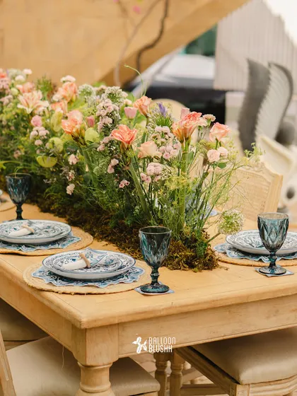 A rustic wooden table set with a lush runner of moss and wildflowers. The blue glassware and patterned plates add a touch of Mediterranean charm.