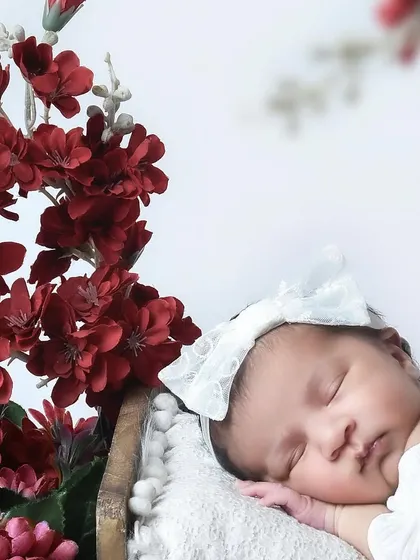A sleeping newborn girl wearing a large white bow, posed next to a beautiful arrangement of deep red flowers. The contrast between the white and red creates a striking image.