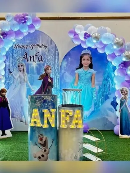 This Frozen theme decor features a personalized backdrop with the birthday girl's photo alongside Anna and Elsa. The setup is completed with a balloon arch, marquee number '6', and character stands.