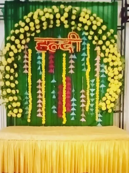 A Haldi backdrop with a circular arch of yellow flowers against a green curtain. The decor includes hanging paper craft for a simple, budget-friendly design.
