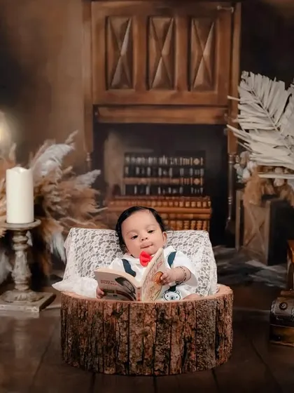 A tiny bookworm reading a story. This cozy, rustic setup with a fireplace and books creates a warm and imaginative setting for a baby photoshoot.