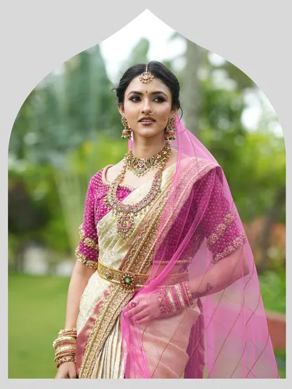 A classic South Indian bridal portrait. The bride is adorned in a cream and gold silk saree with a vibrant magenta blouse, complete with traditional jewelry and a sheer pink veil.