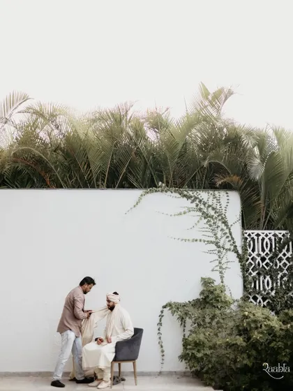 A wide shot of the groom getting his turban tied, set against a clean white wall and lush palm trees, creating a minimalist and stylish portrait.