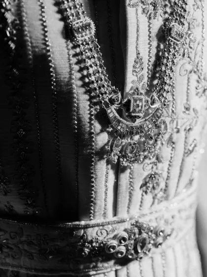 A detailed, artistic black and white shot of the groom's sherwani and necklace. This close-up highlights the intricate craftsmanship and adds a touch of timeless elegance to the wedding album.
