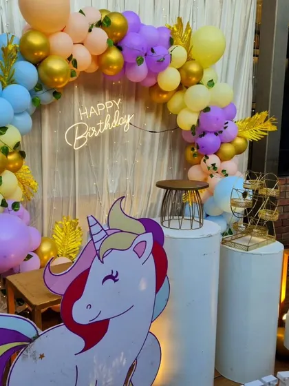 A cheerful unicorn-themed party for a 4th birthday. The setup features a large unicorn cutout, a rainbow-colored balloon arch, and a light-up number.