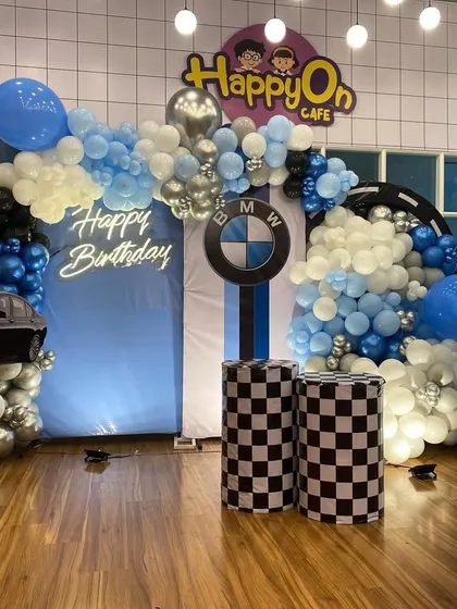 A fun and dynamic BMW car-themed birthday decoration for a 5th birthday. The setup includes a balloon arch in blue, black, and silver, checkered racing flag pedestals, a car cutout, and the iconic BMW logo for a young car enthusiast.