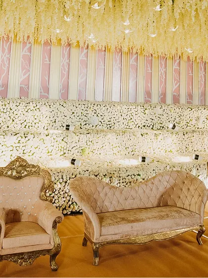 An elegant stage setup featuring a backdrop wall of white flowers and a ceiling of hanging yellow floral strings. The plush, vintage-style seating adds a touch of classic luxury.