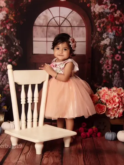 A sweet portrait of a one-year-old standing by a chair in a room filled with flowers and warm light.