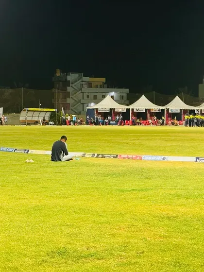 A quiet moment on the boundary during a night game, with the well-lit field and player tents in the background.