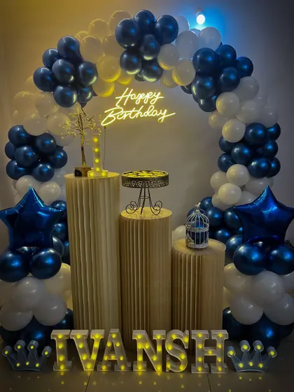 A cool-toned birthday decoration with a blue and white balloon arch, star-shaped foil balloons, and a 'Happy Birthday' neon sign.