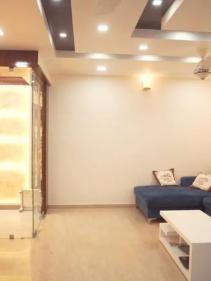 A spacious living room with a large blue sectional sofa and a 3D cushioned wall panel. The false ceiling with dark wood accents and multiple light sources creates a dramatic and modern effect.