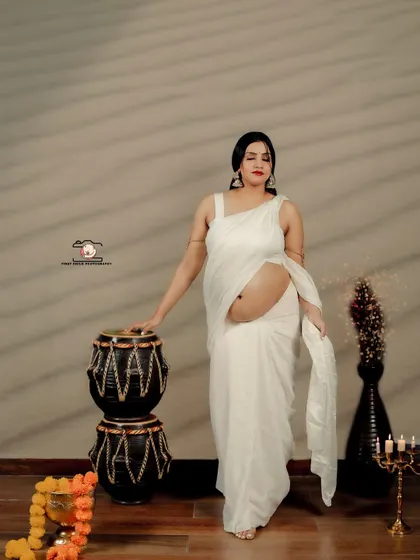 A full-length standing portrait of the mother-to-be in a white saree. The simplicity of the attire is contrasted with traditional props like the tabla and candelabra, creating a unique blend of styles.