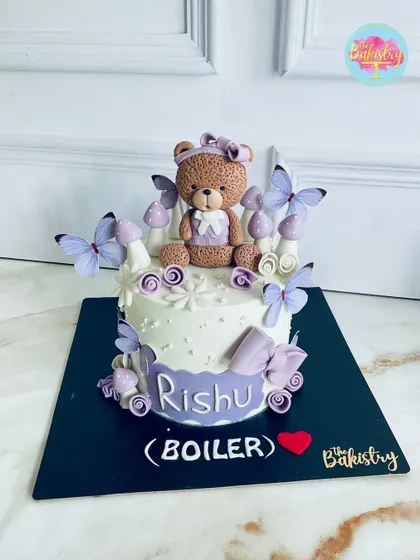 An adorable teddy bear cake in a soft purple color scheme for Rishu's first birthday, complete with whimsical mushrooms and butterflies.