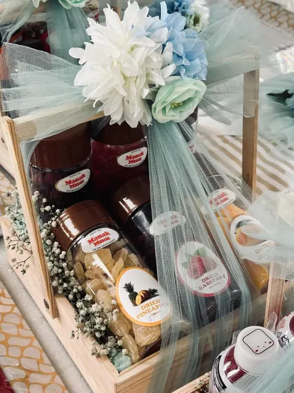 A hamper filled with gourmet dried fruits and other snacks. The clear jars and bottles are beautifully arranged in a wooden tray with floral decorations.