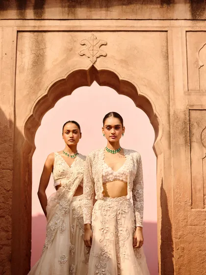 A shot of two models in complementary ivory ensembles, one in a saree and the other in a lehenga with a jacket. This showcases how different styles from one collection can be coordinated for a wedding party.