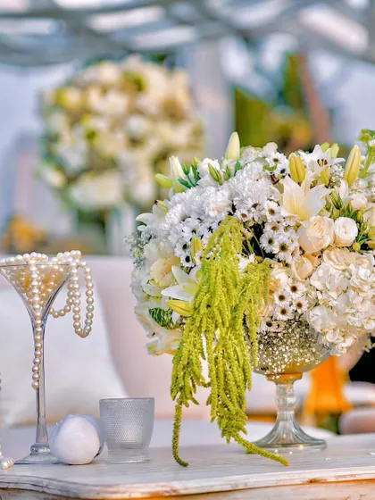 A close-up of the table decor, where a martini glass filled with pearls sits beside a lush arrangement of white lilies, roses, and hanging amaranthus. The details are both chic and playful.