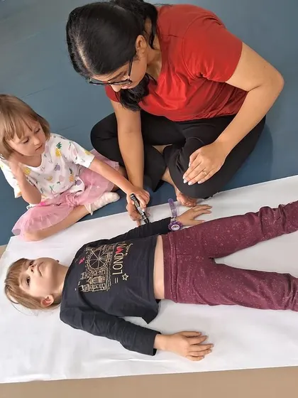 A teacher helps a child trace their body outline on a large sheet of paper, the first step in our "All About Me" anatomy project.