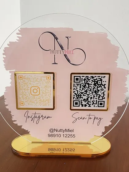 A straight-on view of the 'NuttyMiel' QR stand, showcasing the clear acrylic and the elegant contrast between the pink, black, and gold elements.