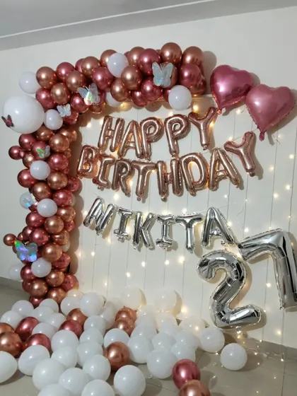 A 27th birthday home decoration with a rose gold and white balloon arch, 'Happy Birthday' foil letters, and a fairy light curtain.