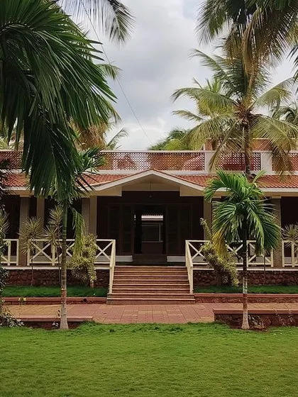 The symmetrical design of the 'Grid in the Grove' farmhouse is evident from this frontal view. The building is perfectly centered within the lush green lawn and surrounding palm trees.
