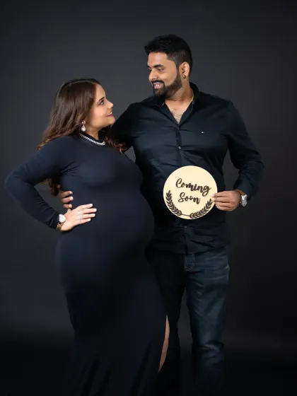A stylish maternity announcement photo. The couple, dressed in black, holds a 'Coming Soon' sign, creating a chic and modern portrait against a dark backdrop.