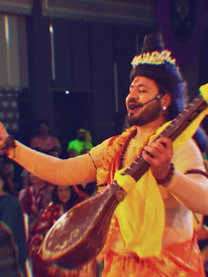 Performing as Narada Muni with a veena in hand. This isn't just hosting; it's a full performance that requires acting, singing, and engaging the audience in character.