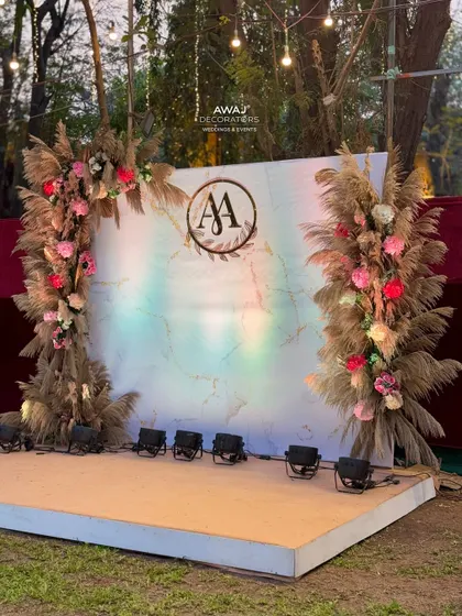 A beautiful boho-chic stage for an outdoor sangeet, with pampas grass arches, a marble-effect backdrop, and a custom monogram.