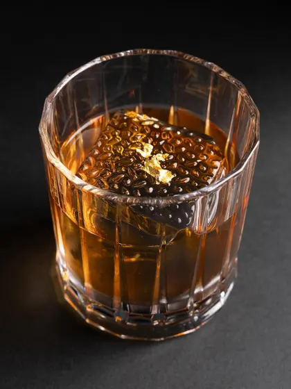 A close-up of a whiskey cocktail with a unique, textured ice cube and a flake of gold leaf, emphasizing luxury and detail.