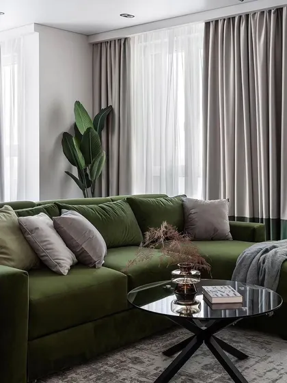 A living room featuring a plush olive green L-shaped sofa, layered curtains, and natural elements. The design creates a comfortable and earthy vibe.