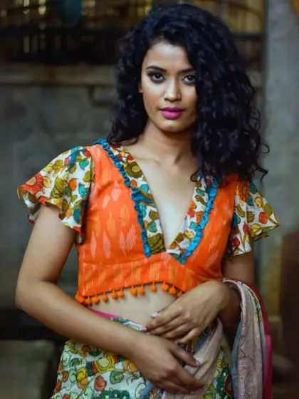 A vibrant orange Kalamkari blouse with a deep V-neck and playful flutter sleeves. The pom-pom tassels add a fun, bohemian touch to the traditional print.
