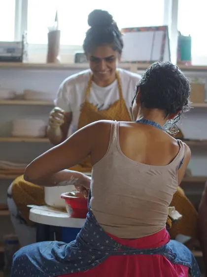 A beautiful shot of two students working at their wheels, with one smiling in the background, capturing the supportive and happy atmosphere of the studio.