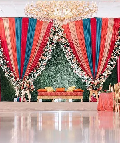 A vibrant Sangeet stage with colorful draping and a green turf backdrop. The mix of teal, orange, and red fabrics creates a festive and energetic atmosphere.