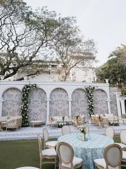 An elegant outdoor lounge area with a European feel, featuring a white arched backdrop with delicate floral patterns, comfortable seating, and classic decor elements.