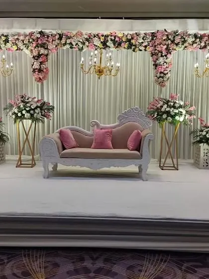 A beautiful pastel-themed stage. A classic white sofa is framed by floral arrangements on pedestals, with a backdrop of white curtains and hanging chandeliers creating a soft, dreamy look.