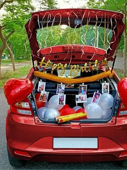 A sweet and simple car trunk birthday surprise. The car boot is filled with balloons, a 'Happy Birthday' banner, and a string of memorable photos.