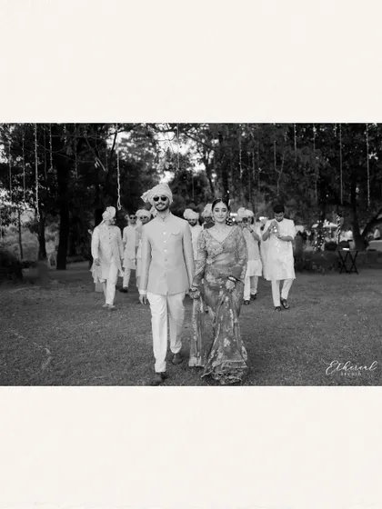 A black and white photograph of the bride and groom walking with their entourage, yet they are the clear focus. It captures the "marriage of equals" concept with a timeless, cinematic feel.
