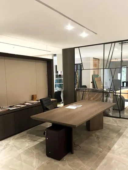This image captures an office during the final stages of furnishing. A large, minimalist wooden desk is in place, with custom dark wood cabinetry along the wall. The glass partition with a geometric black frame separates the space while maintaining an open feel.