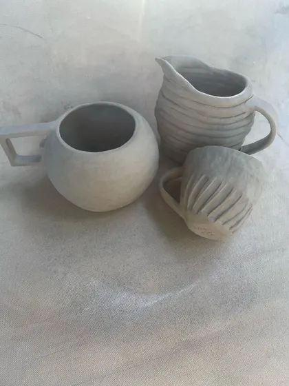 A set of unfired hand-built mugs and a pitcher. Each piece has a unique shape and texture, from the smooth, round mug to the carved and fluted smaller cup.