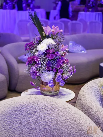 A beautiful floral arrangement of purple and white flowers in a gold vase, placed in a lounge area with plush, modern seating under purple lighting.