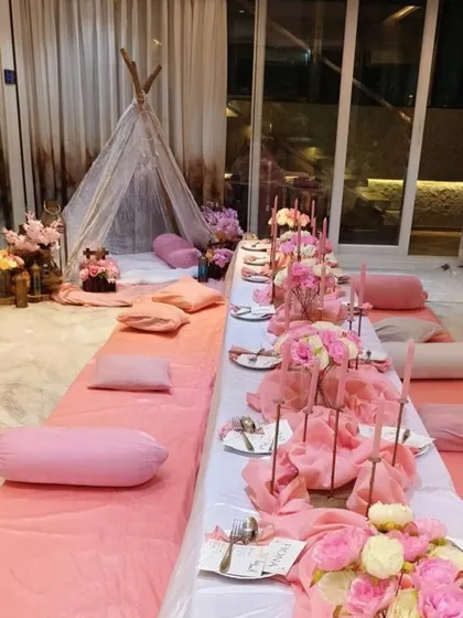A cute and cozy sit down dinner setup, all in shades of pink. This low seating arrangement features pink floor coverings and cushions, a long table with pink floral centerpieces, and a lace teepee tent in the corner for a whimsical touch.