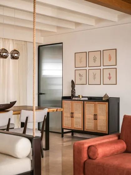 The open-plan living and dining area is zoned diagonally to create flow and openness. A cane-fronted credenza provides storage and adds a touch of natural texture, complemented by botanical art prints.