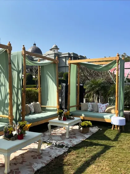 A relaxed outdoor lounge area with canopy beds draped in pastel green fabric, perfect for a sunny afternoon Mehendi or Haldi event.