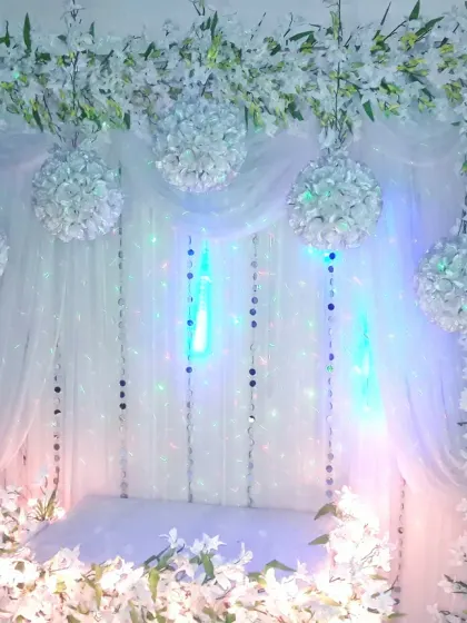 An elegant all-white decoration for a home festival or pooja. The backdrop is framed with white flowers and features hanging floral balls and subtle mood lighting.