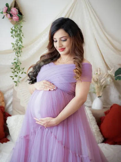 A serene moment in a cozy, boho-inspired setting. The mom-to-be cradles her bump, dressed in a lovely lavender tulle gown.