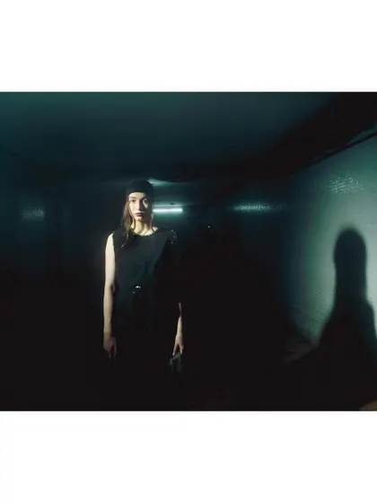 A wide shot of a model in a dark, narrow hallway, with her shadow cast starkly against the wall. This image uses minimal light to create a cinematic, noir-inspired atmosphere of suspense and solitude.