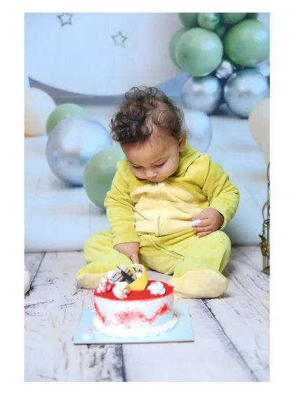 A baby in a dinosaur onesie curiously inspecting his first birthday cake.